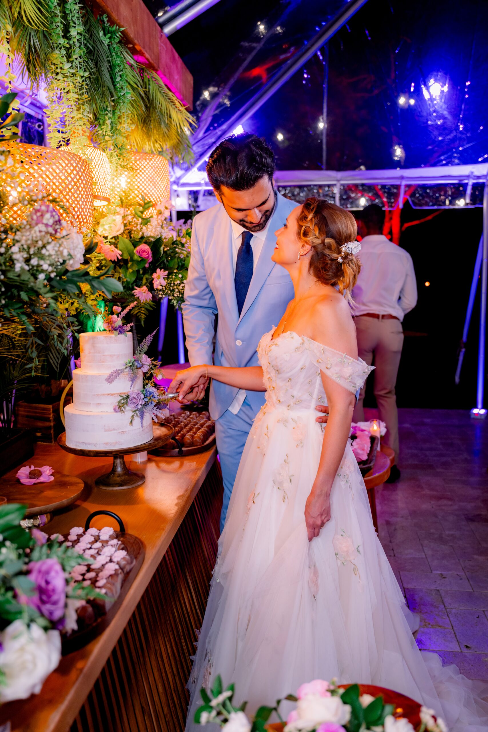 Couple cutting a floral wedding cake under a clear tent with lavender décor at Saint Patrick Palace in Davie, FL.