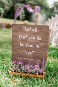 Wooden ceremony sign with lavender florals and a Bible verse at a lakeside wedding at Saint Patrick Palace.