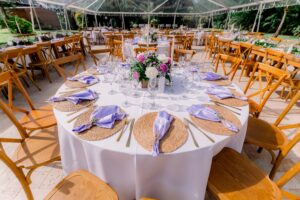 Round wedding table with woven chargers, lavender napkins, and floral centerpiece under a clear tent.