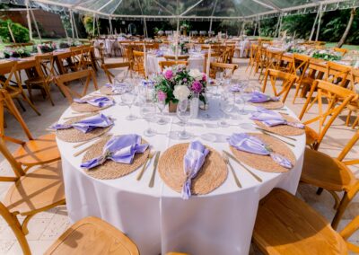 Round wedding table with woven chargers, lavender napkins, and floral centerpiece under a clear tent.