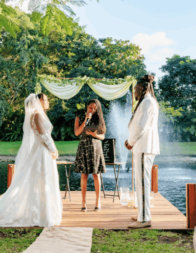 Bride and groom exchange vows on a lakeside dock with greenery and fountain backdrop at a private estate wedding.