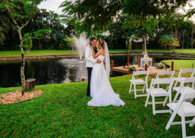 Bride and groom share a romantic kiss at a lakeside wedding ceremony with fountain backdrop.