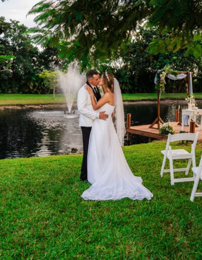Bride and groom share a romantic kiss at a lakeside wedding ceremony with fountain backdrop.