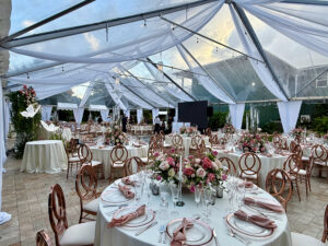 Rose-gold quinceañera celebration under clear tent with chandelier and florals.