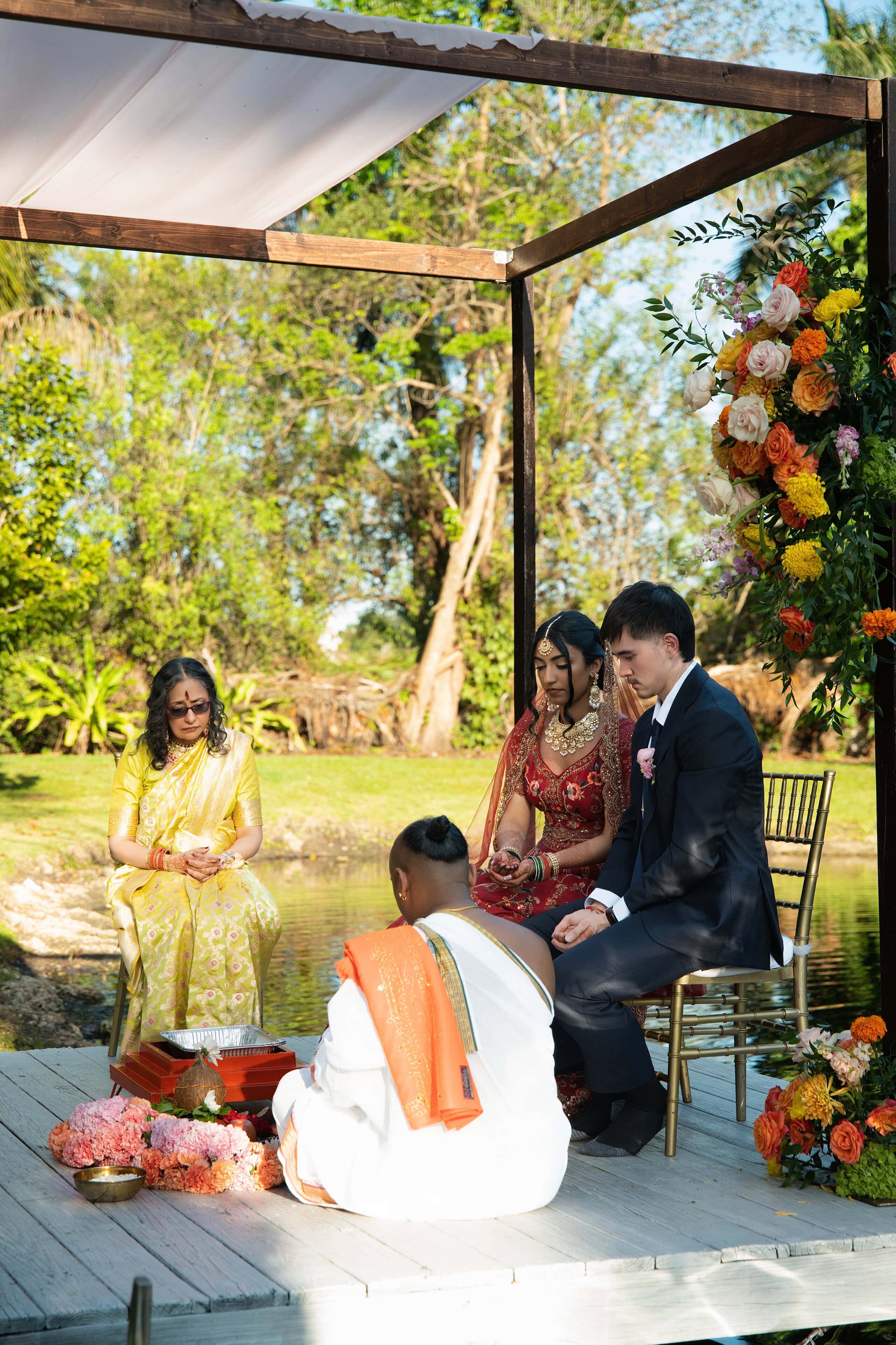 Couple seated under mandap during traditional ceremony at Saint Patrick Palace with officiant and family by the lake.