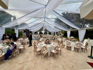 Elegant quinceañera reception under clear tent with draped ceiling and rose-gold décor.