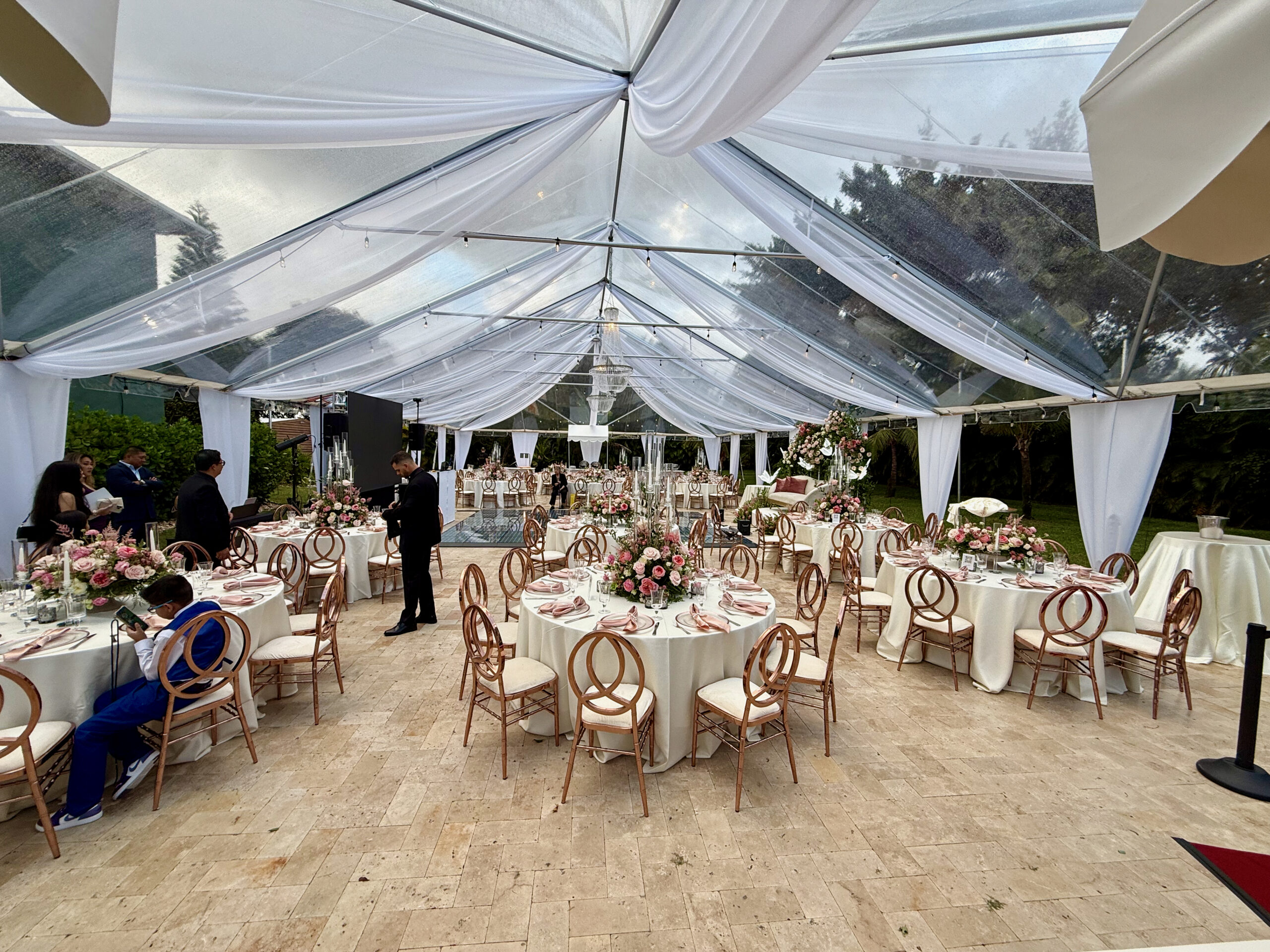 Elegant quinceañera reception under clear tent with draped ceiling and rose-gold décor.