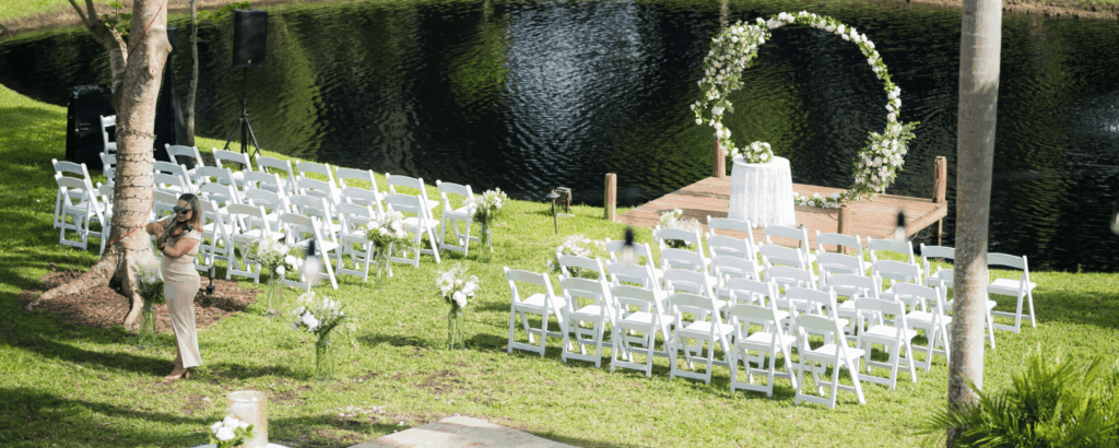 Wide shot of a peaceful outdoor ceremony with white chairs and a circular floral arch on a wooden dock over the water.